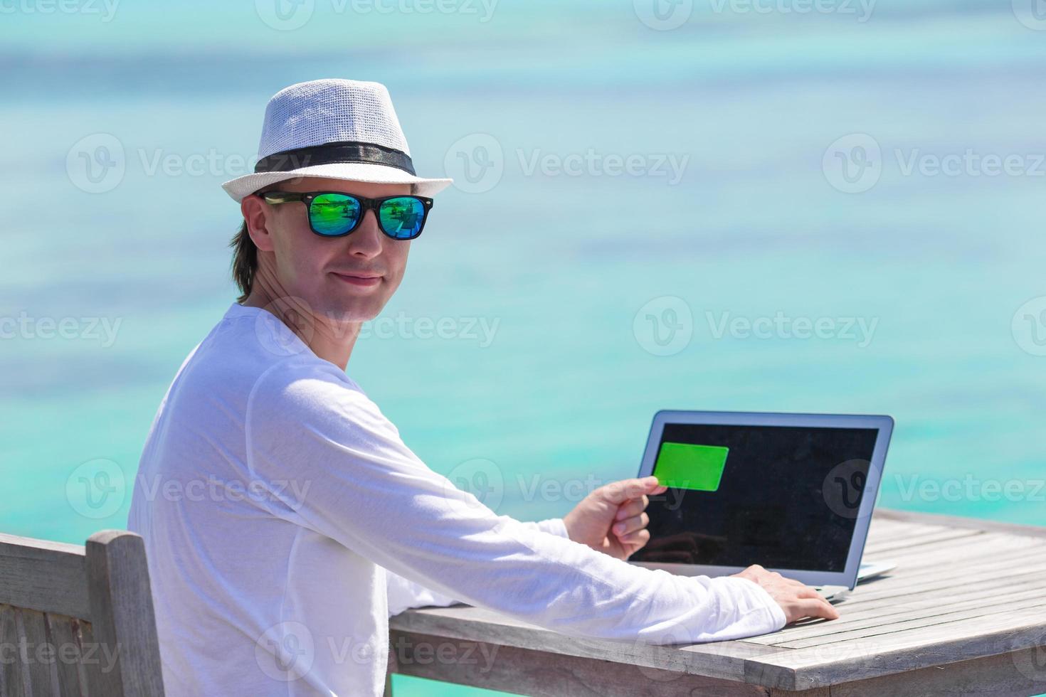 Young man working on laptop with credit card at tropical beach photo