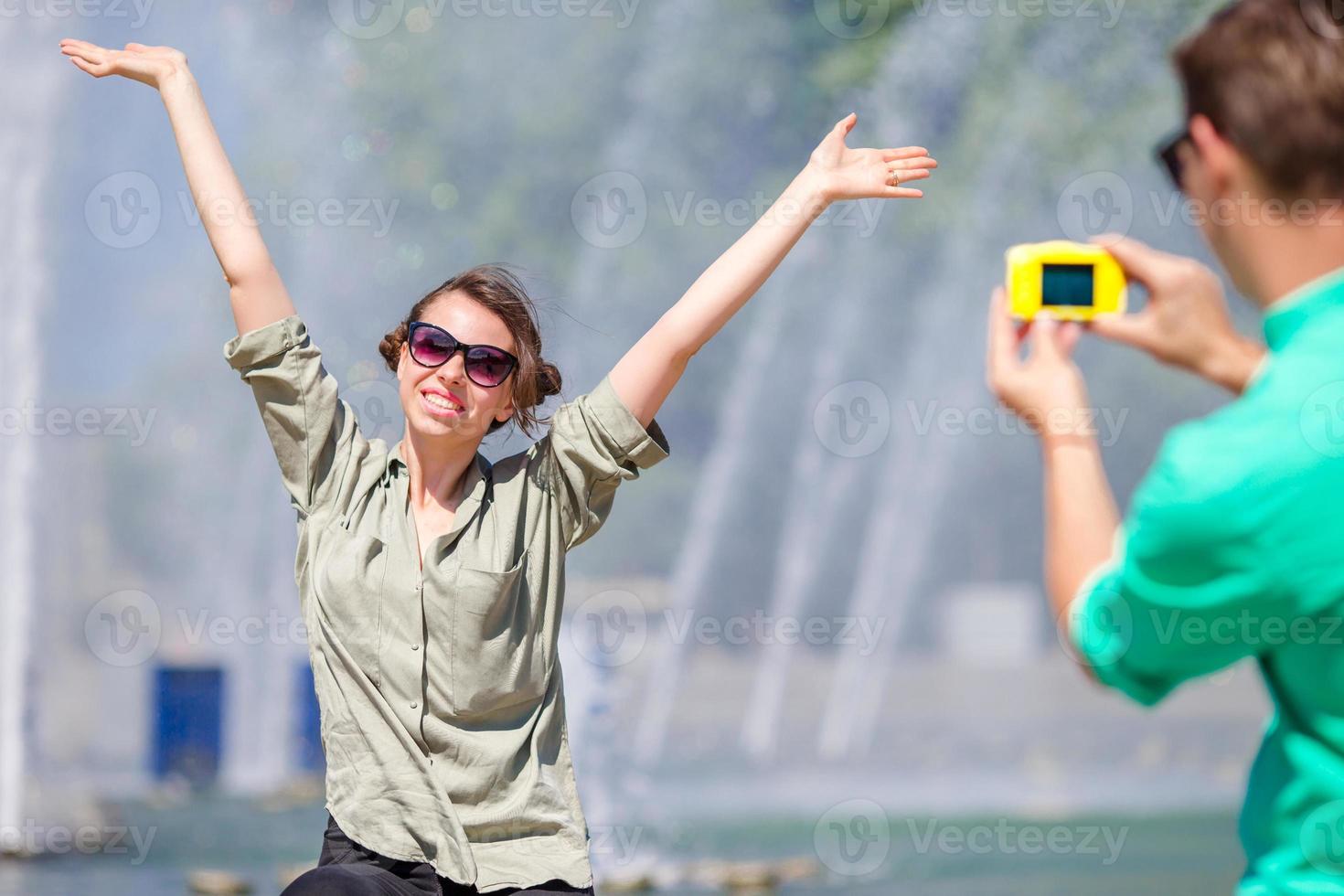 novio tomando una foto de su novia mientras se sienta al fondo de la fuente. joven haciendo fotos de mujer en la calle riendo y divirtiéndose en verano.