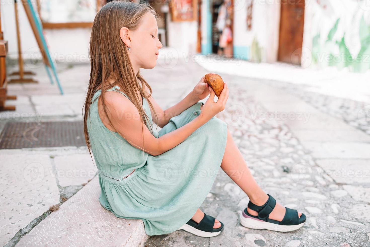 Little girl eats croissant on old streets in european village photo