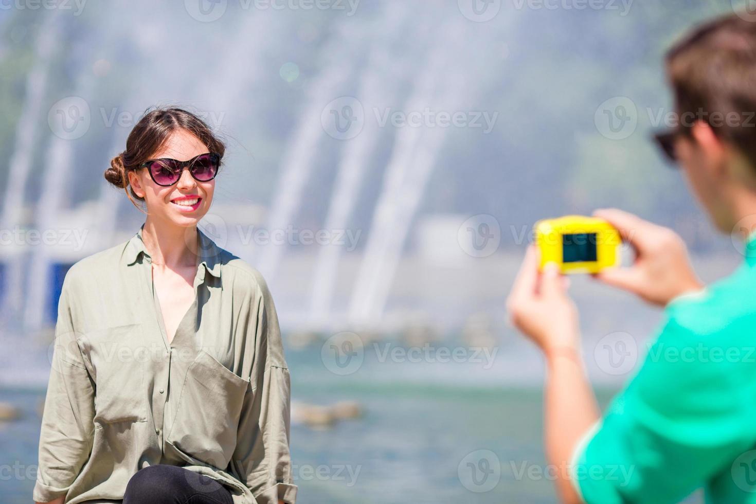 novio tomando una foto de su amigo mientras está sentado al fondo de la fuente. joven haciendo fotos de mujer en la calle riendo y divirtiéndose en verano.