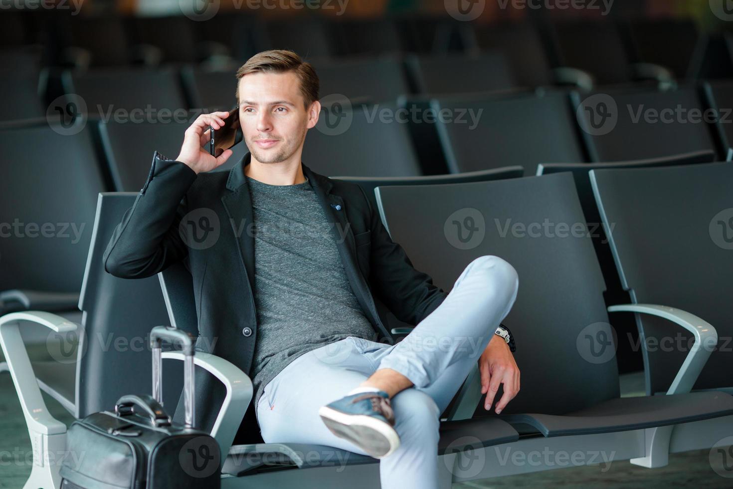 hombre de negocios urbano hablando por teléfono inteligente viajando dentro del aeropuerto. hombre de negocios joven casual con chaqueta de traje. guapo modelo masculino. joven con celular en el aeropuerto mientras espera el embarque. foto