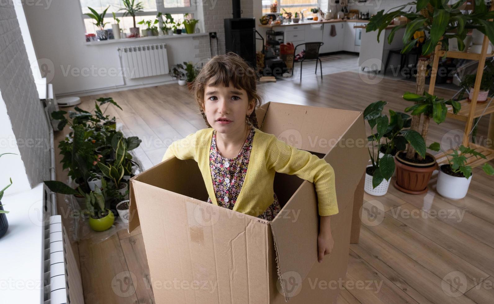 The girl hides in a big box and looks out of it funny in a room inside the home. Moving to a new house, unpacking things, renting an apartment, mortgage, housing issue photo