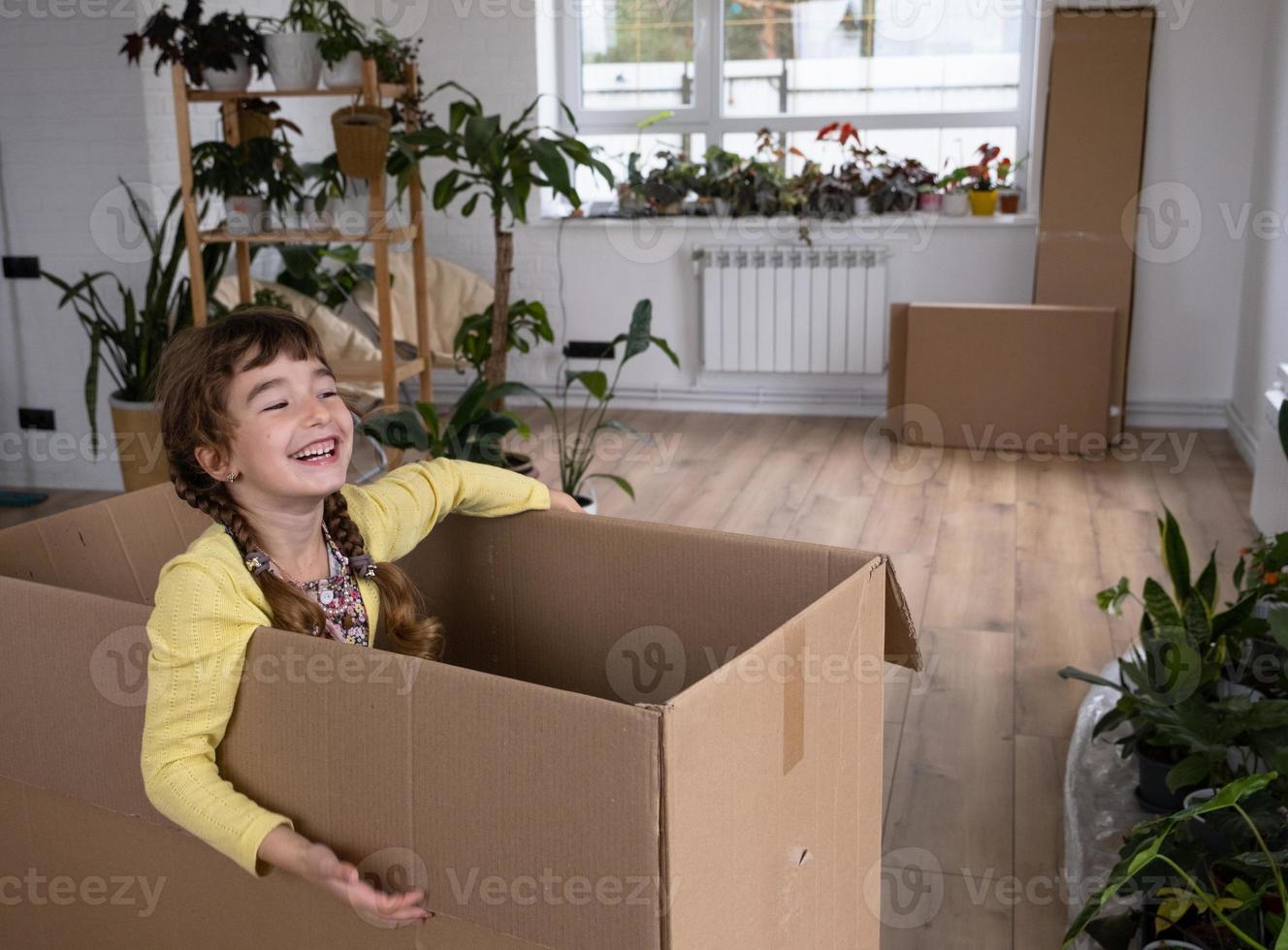 la niña se esconde en una caja grande y mira fuera de ella divertida en una habitación dentro de la casa. mudarse a una casa nueva, desempacar cosas, alquilar un apartamento, hipoteca, problema de vivienda foto