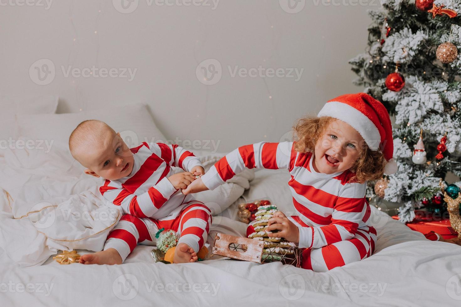 los niños en pijama rojo y blanco comen dulces navideños sentados en la cama. hermano y hermana, niño y niña comparten regalos. mañana de Navidad. estilo de vida. espacio para texto. foto de alta calidad