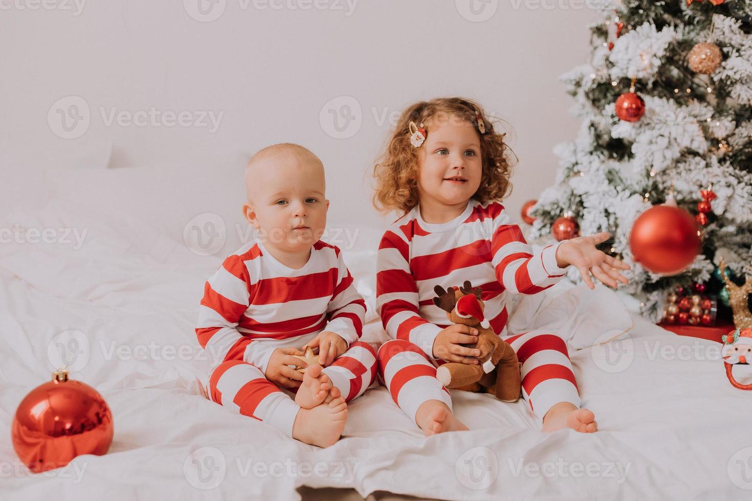 los niños en pijama rojo y blanco comen dulces navideños sentados en la cama. hermano y hermana, niño y niña comparten regalos. mañana de Navidad. estilo de vida. espacio para texto. foto de alta calidad