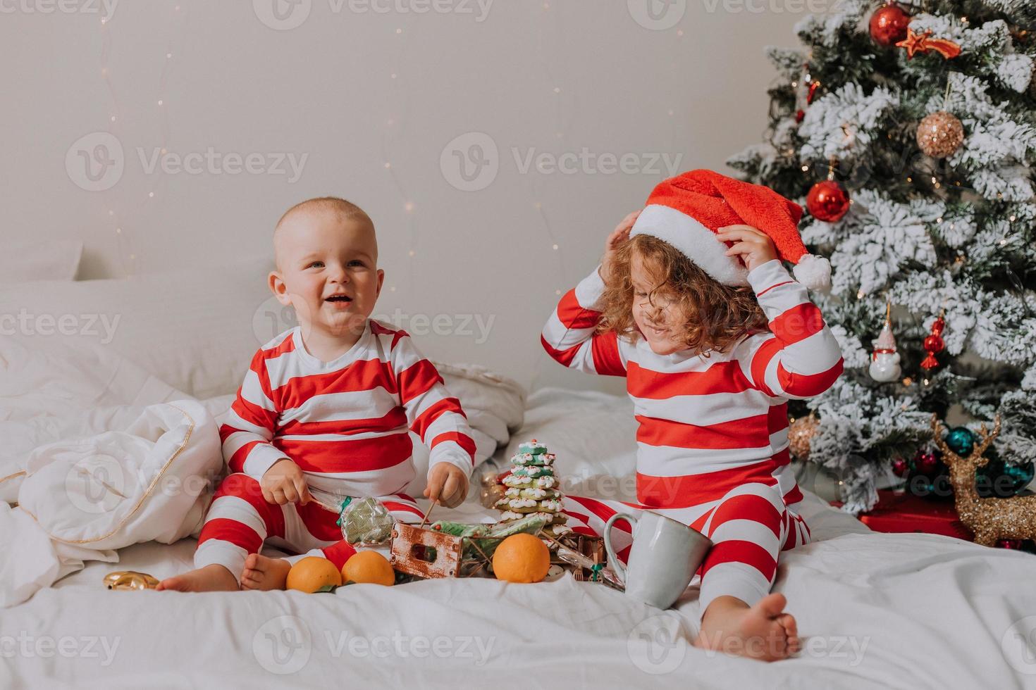 children in red and white pajamas eat Christmas sweets sitting in bed. brother and sister, boy and girl share gifts. christmas morning. lifestyle. space for text. High quality photo