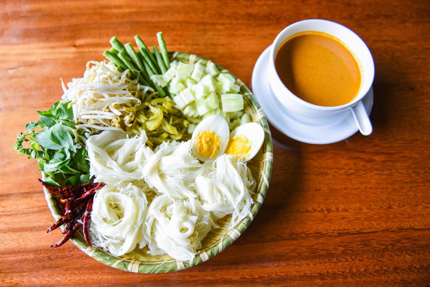 Fideos de arroz tailandeses: cierre la comida de Tailandia fideos vermicelli huevos cocidos y verduras frescas en un plato con un tazón de sopa de curry servido en una mesa de madera foto