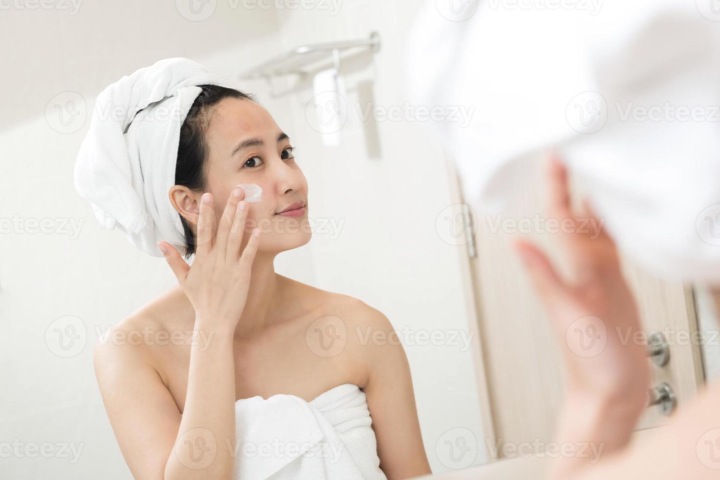 Happy young Asian woman applying face lotions while wearing a towel and touching her face in bathroom photo