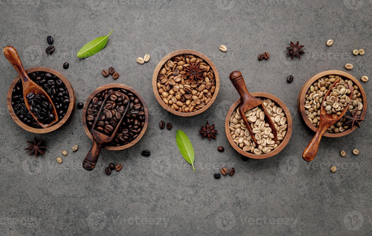 Green and brown unroasted and dark roasted coffee beans in wooden bowl with spoons set up on dark concrete background. photo