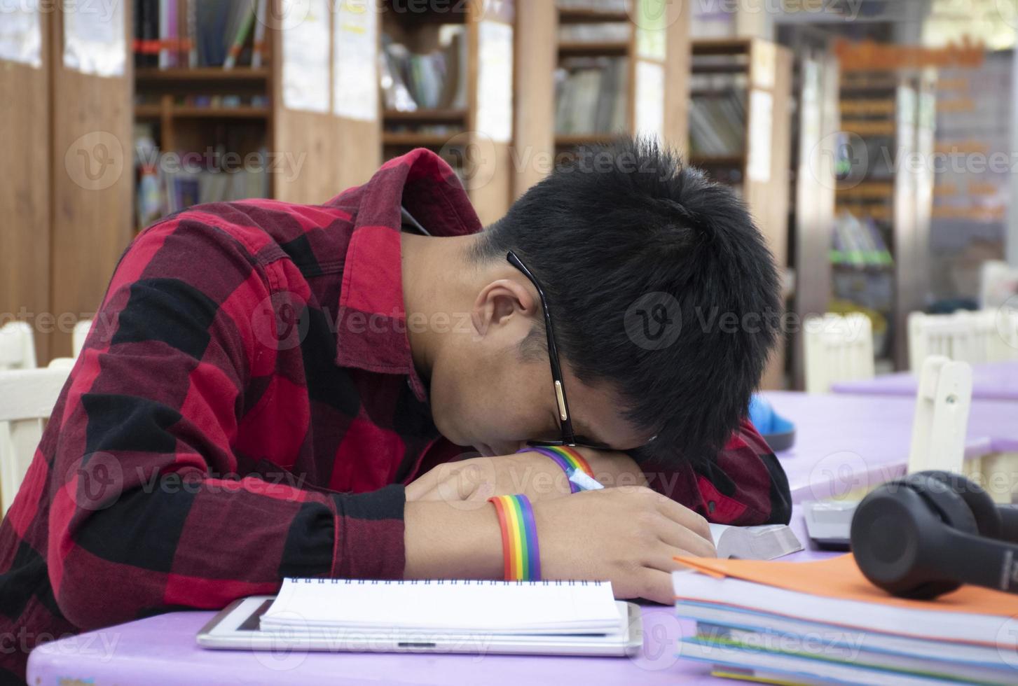 Young Asian boy wears rainbow wristband, sitting in library and taking a nap on table while doing his hard school project work, concept for raising teens and LGBT people daily life activity. photo