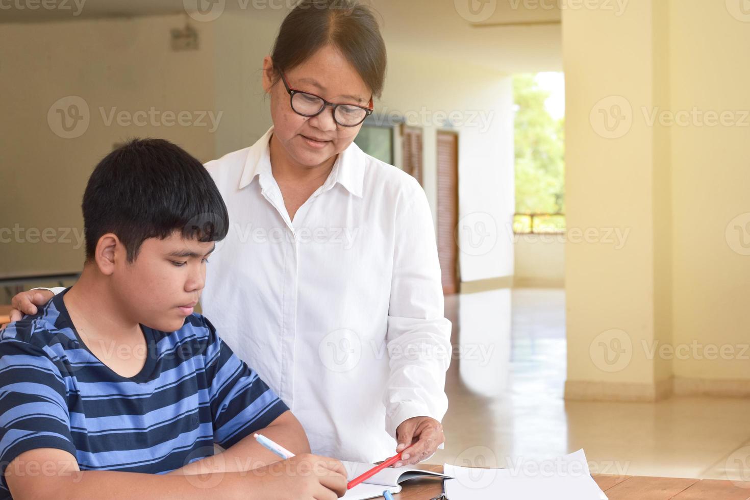 Young asian boy is doing his assignment and listening to his female elderly teacher's advices about school project, adult helps kids to do school project concept. photo