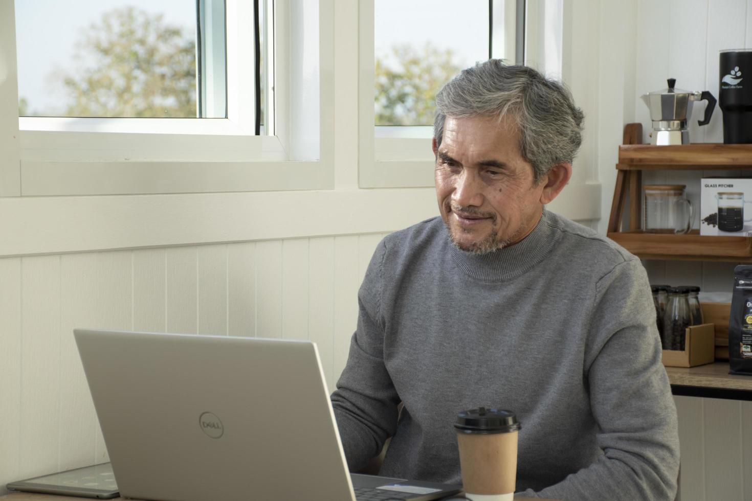 Portrait elderly senior asian man sits near glass window in the morning to work from home and checking his business on his laptop on table seriously, soft and selective focus. photo