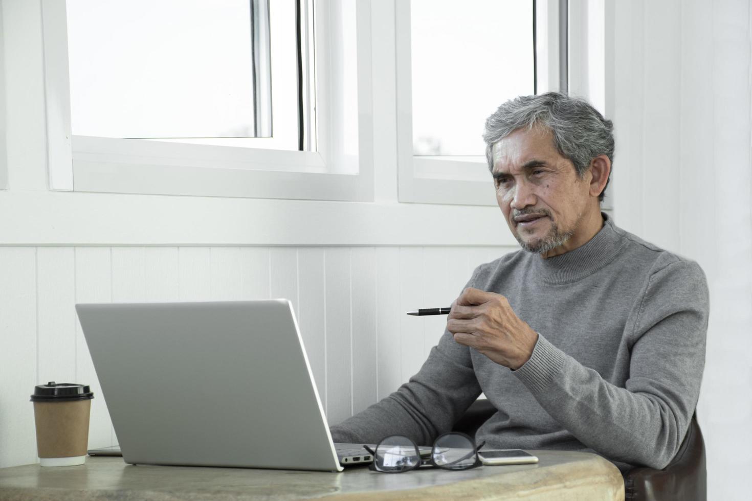 Portrait elderly senior asian man sits near glass window in the morning to work from home and checking his business on his laptop on table seriously, soft and selective focus. photo