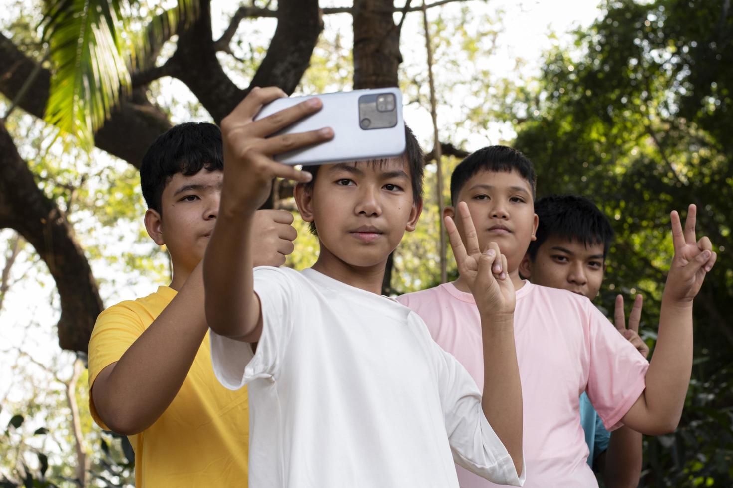 Group of young asian teen boys spending free times in the park rasing their fingers and taking selfie together happily, soft and selective focus on boy in white t-shirt, raising teens concept. photo