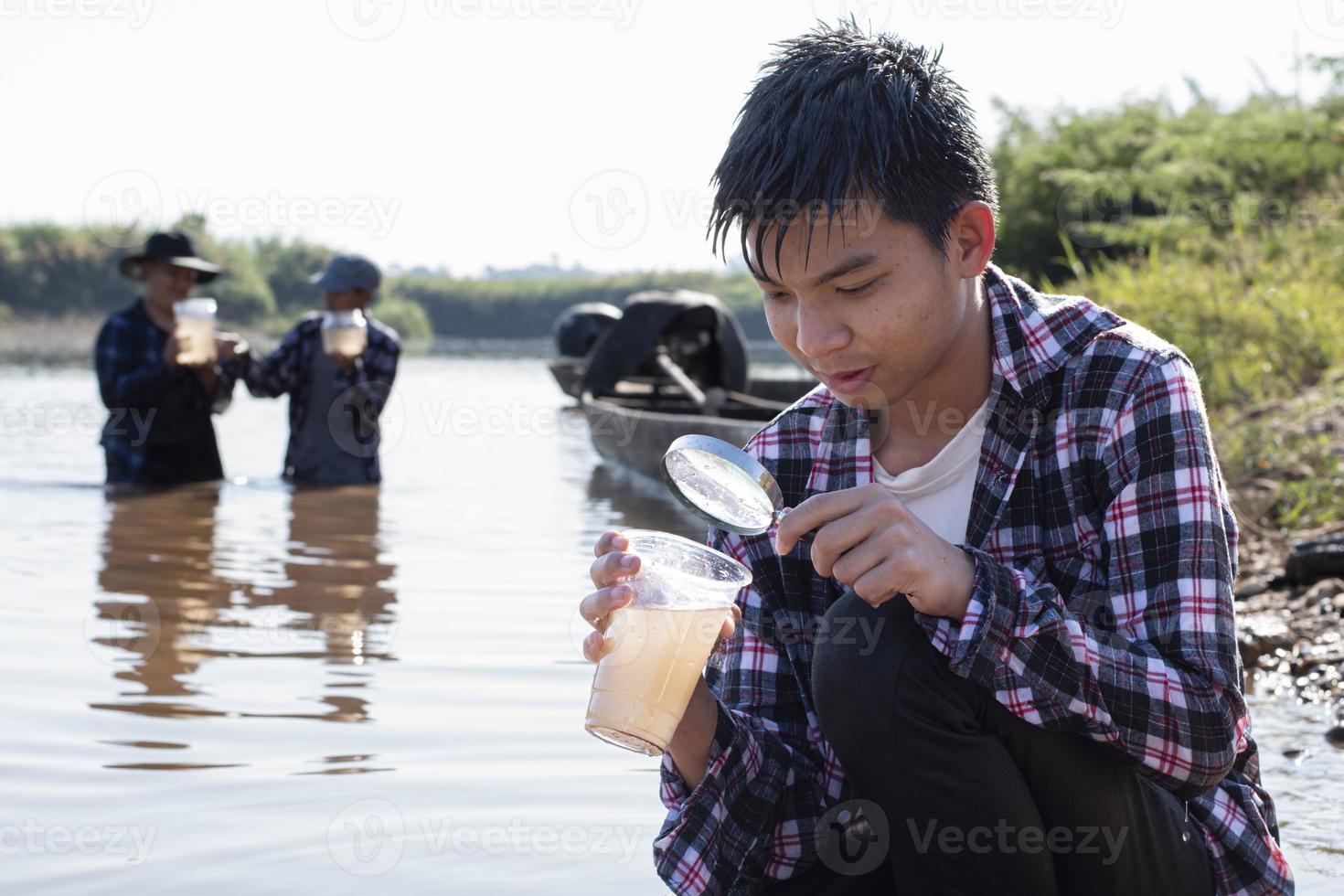 Young asian boy holds transparent tube which has example water inside to do the experiment and ph level measurement as his school project work with his friends behind at the river where he lived. photo