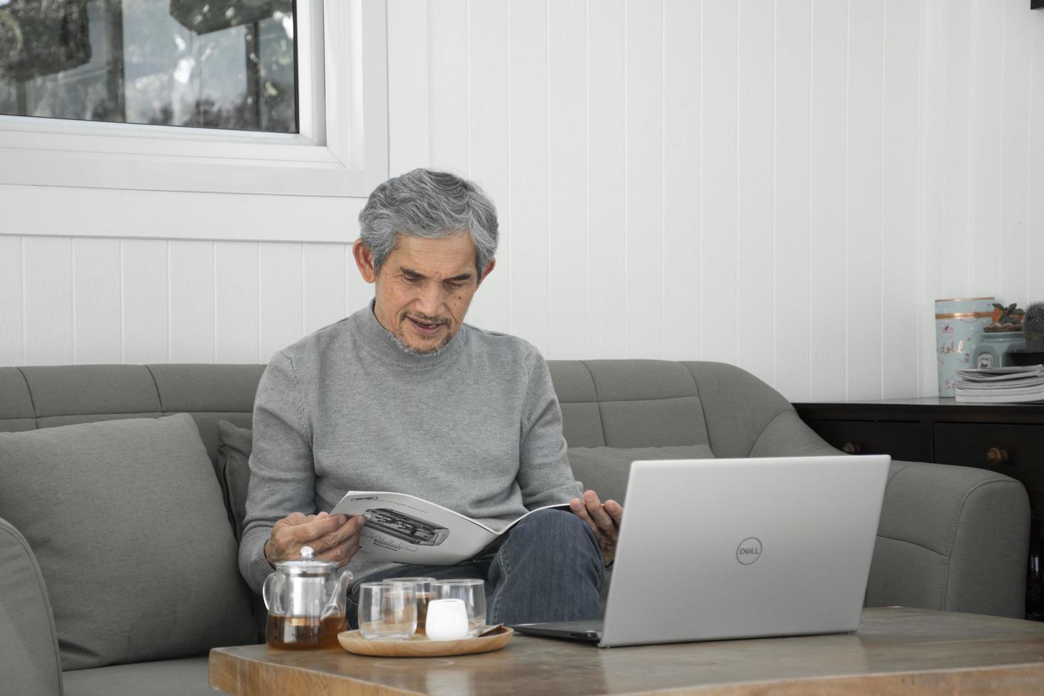 Portrait elderly senior asian man sits near glass window in the morning to work from home and checking his business on his laptop on table seriously, soft and selective focus. photo