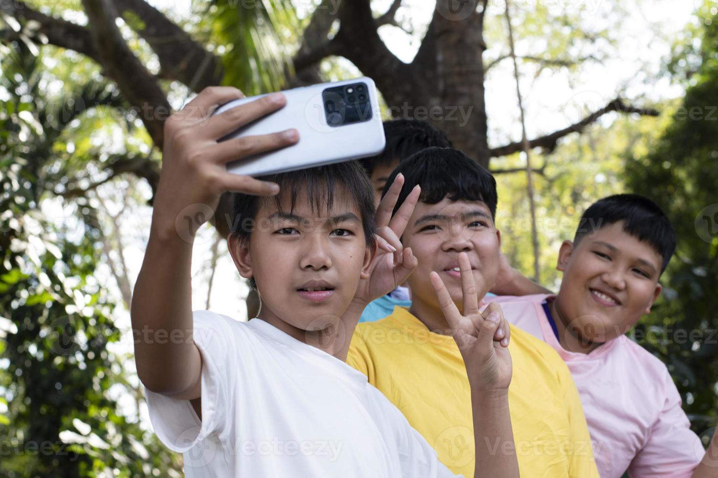 Group of young asian teen boys spending free times in the park rasing their fingers and taking selfie together happily, soft and selective focus on boy in white t-shirt, raising teens concept. photo
