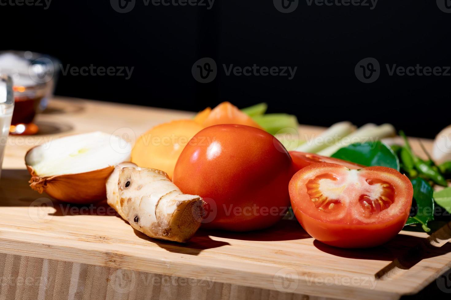 Tomato and sliced half of tomato beside it with onion and galangal, on wood plate in studio light with dark theme. photo