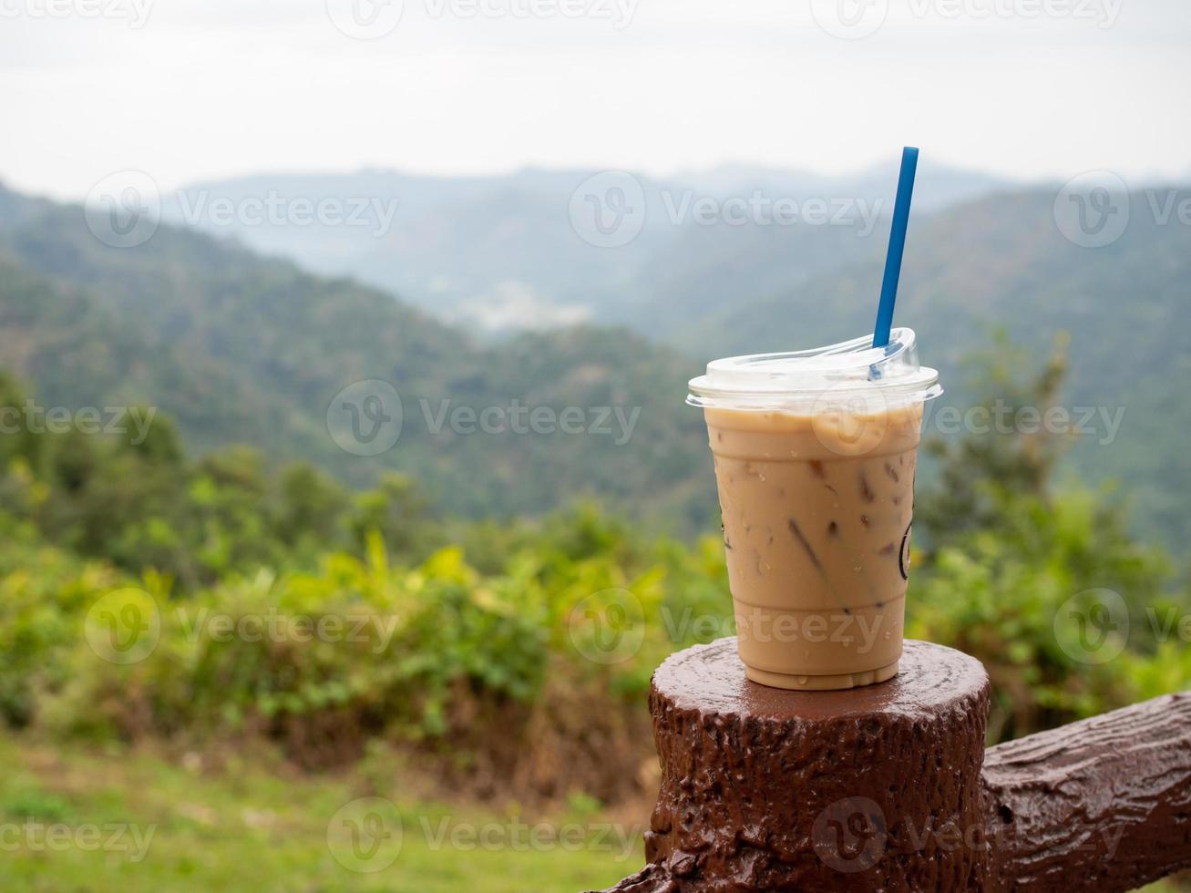 A glass of iced coffee is placed on the fence against a backdrop of mountains and sky. photo