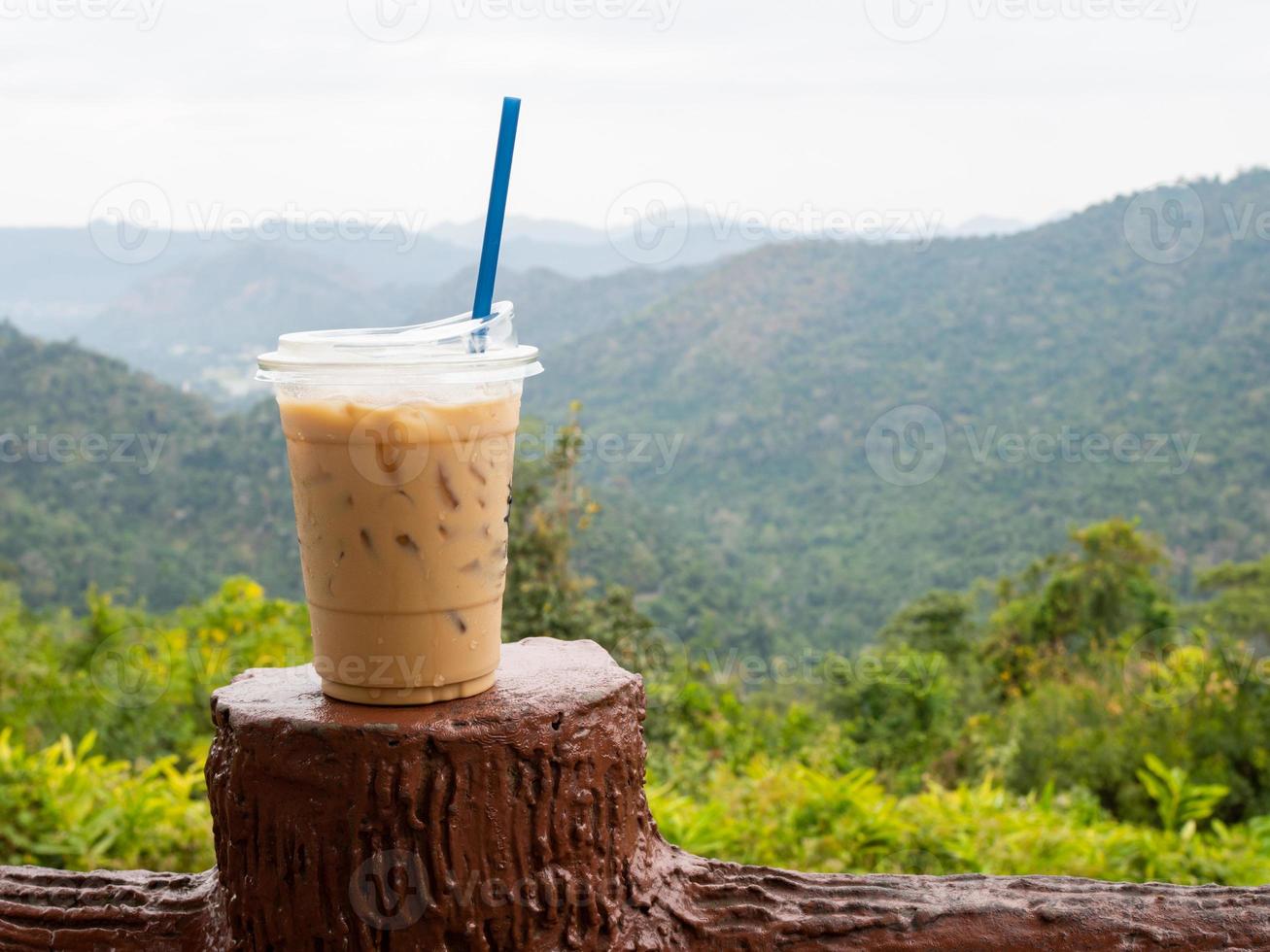 A glass of iced coffee is placed on the fence against a backdrop of mountains and sky. photo