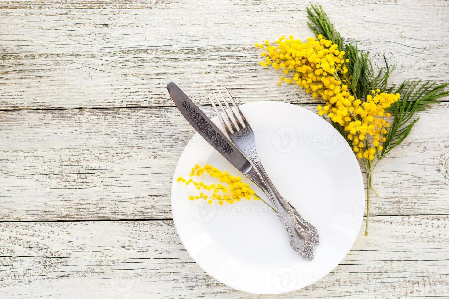 Festive table setting plate with fork and knife and mimosa flower decoration on white wooden background with copy space photo