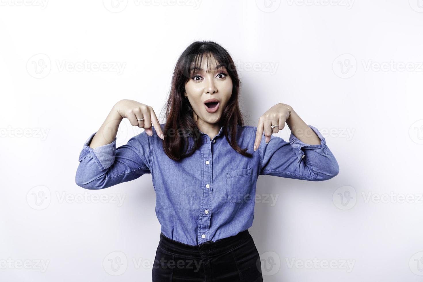 Shocked Asian woman wearing blue shirt pointing at the copy space below her, isolated by white background photo