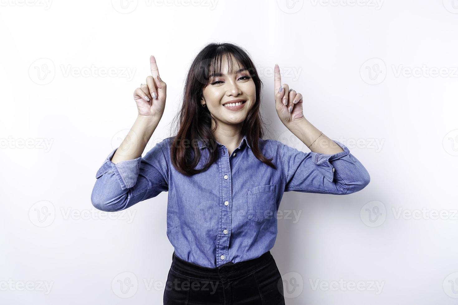 mujer asiática emocionada con pantalones azul apuntando al espacio de copia encima de ella, aislada por fondo blanco foto