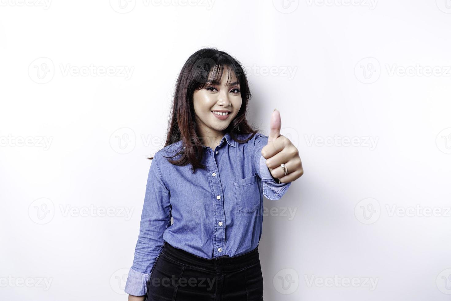 Excited Asian woman wearing blue shirt gives thumbs up hand gesture of approval, isolated by white background photo