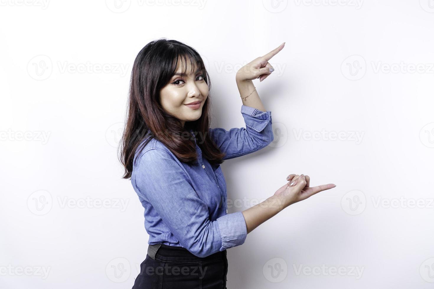 Excited Asian woman wearing blue shirt pointing at the copy space beside her, isolated by white background photo