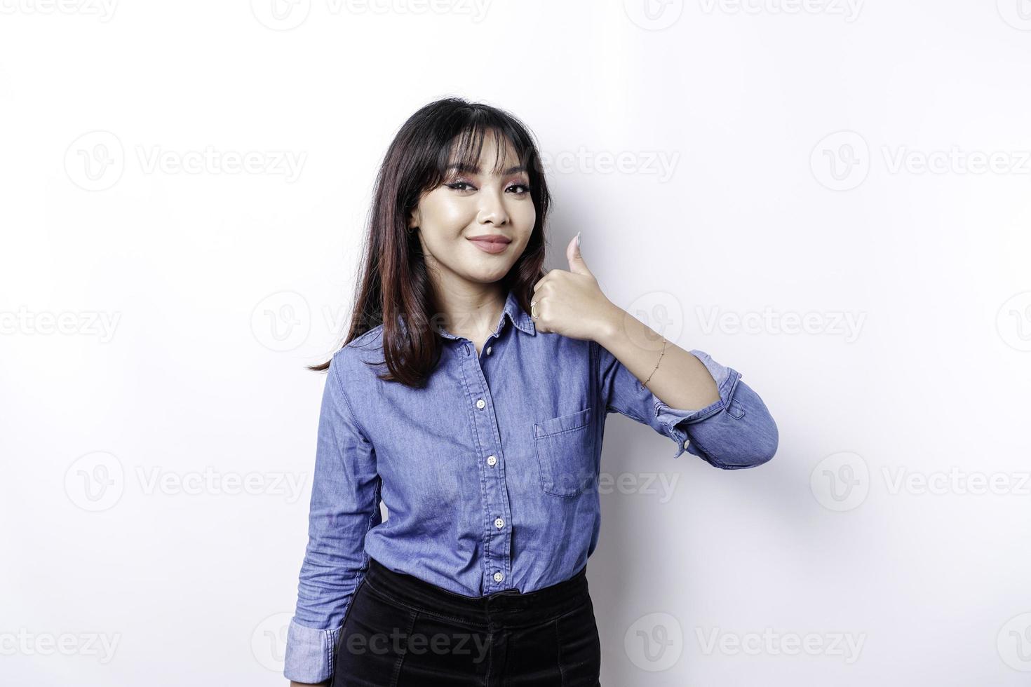 Excited Asian woman wearing blue shirt gives thumbs up hand gesture of approval, isolated by white background photo