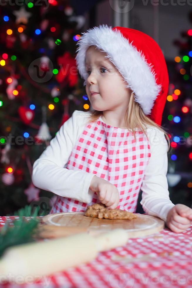 Adorable little girl in Santa hat baking gingerbread Christmas cookies at home photo