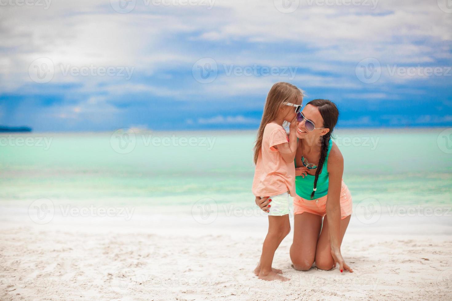 joven madre y su linda niña susurrando en la playa blanca foto