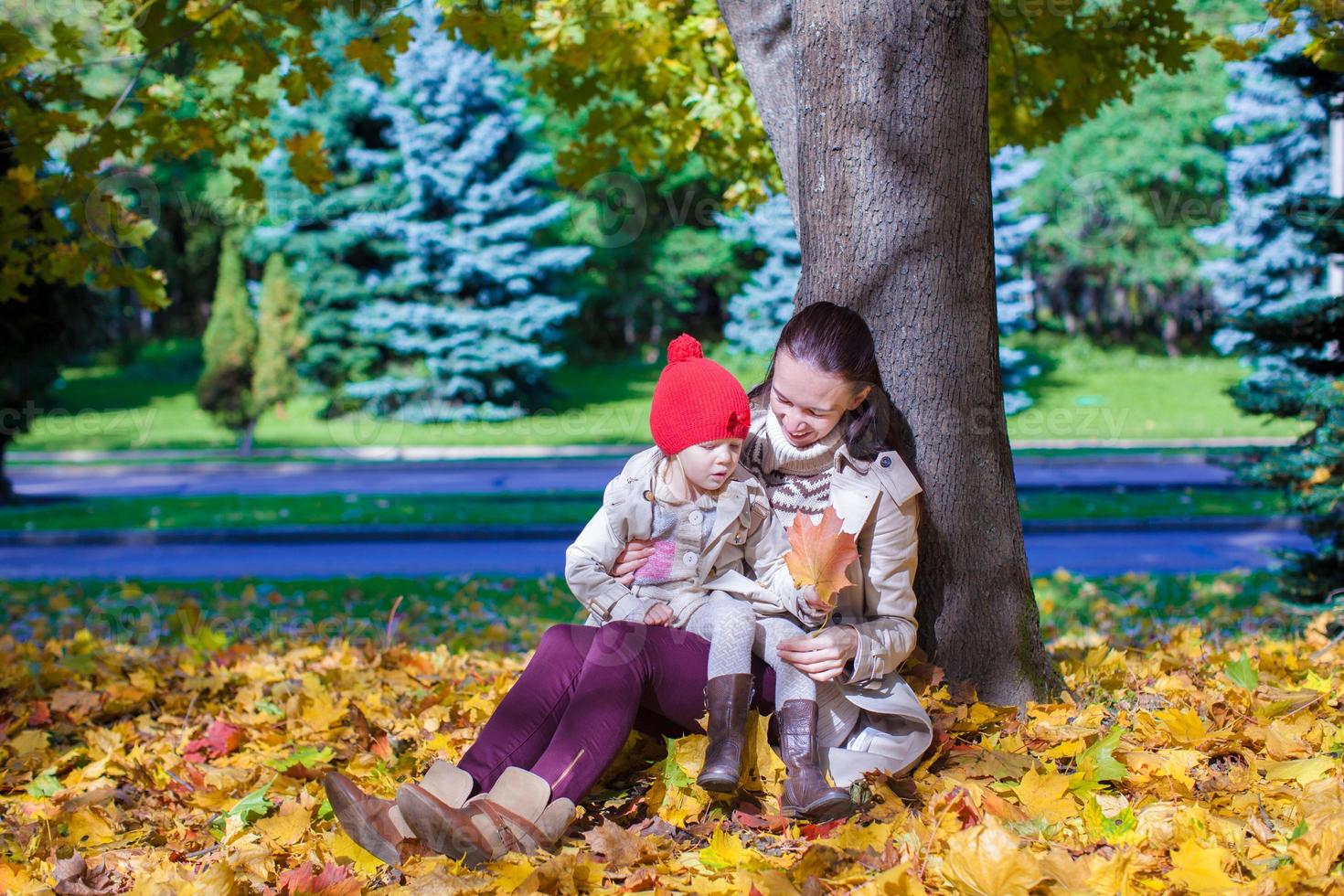 Fashion young mother and her adorable little daughter sitting near big maple in beautiful autumn day photo