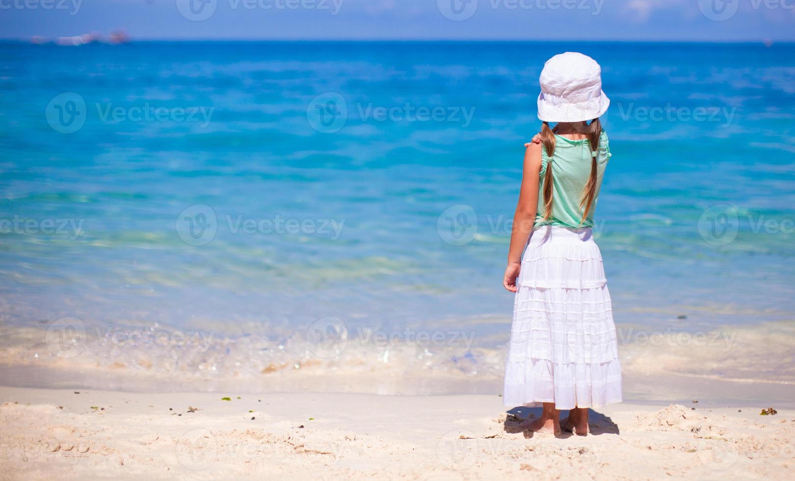 vista trasera de una niña con sombrero mirando el mar en una playa de arena blanca foto