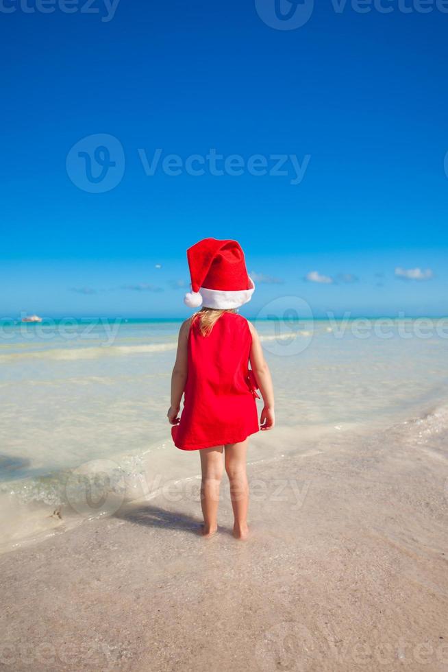 Back view of Little cute girl in red hat santa claus on the beach photo