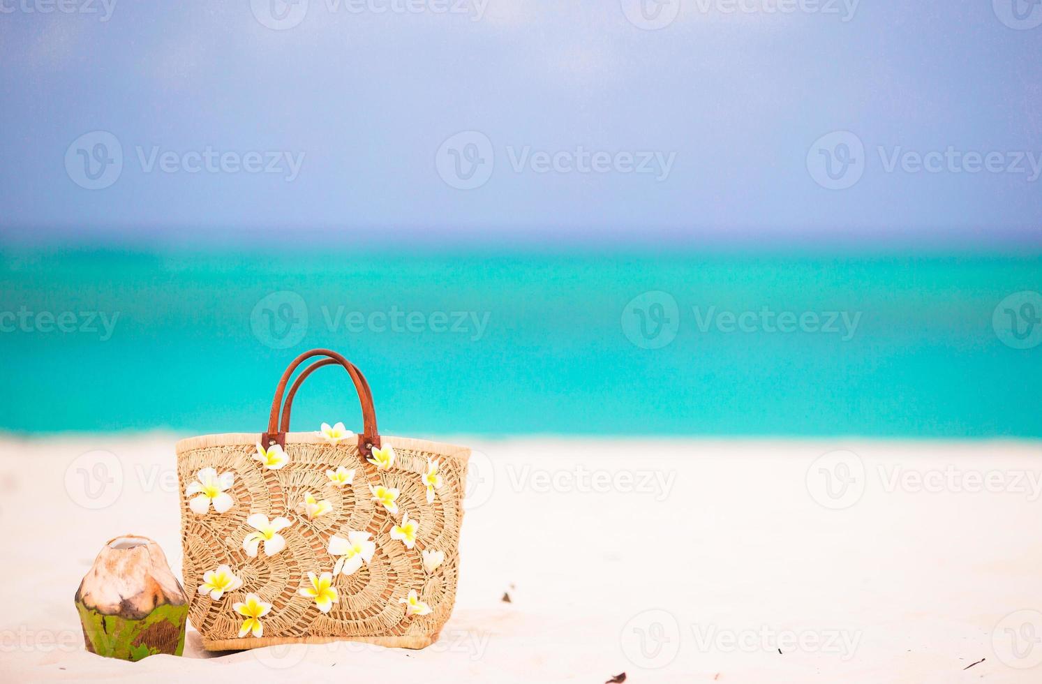 Beach bag with frangipani flowers and coconut on white beach. Closeup beach accessories. photo