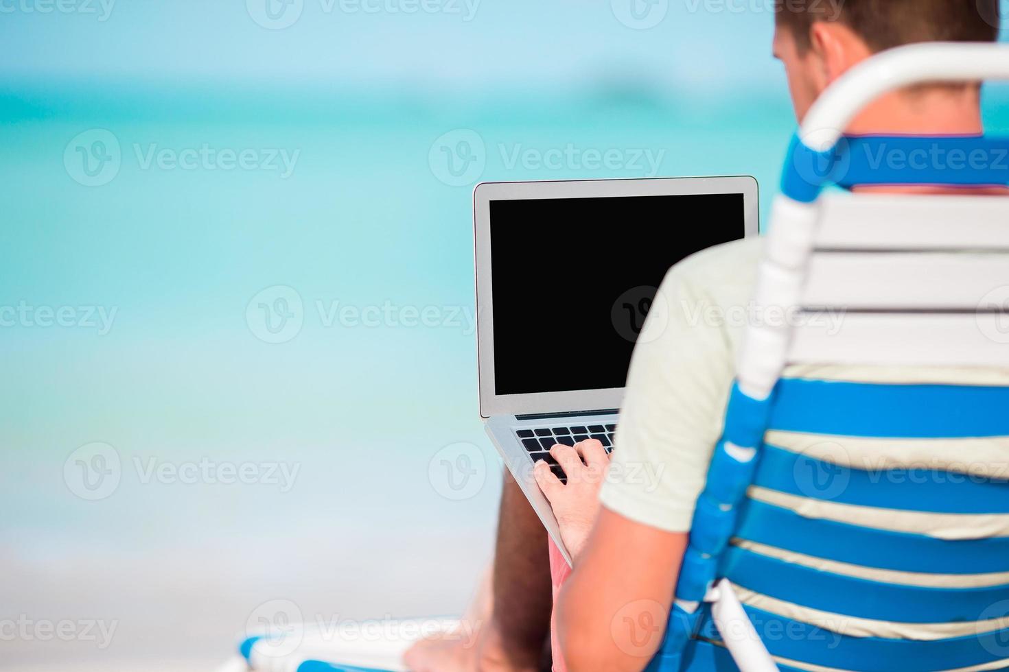 Young man with laptop on tropical caribbean beach. Close up computer in male hands photo