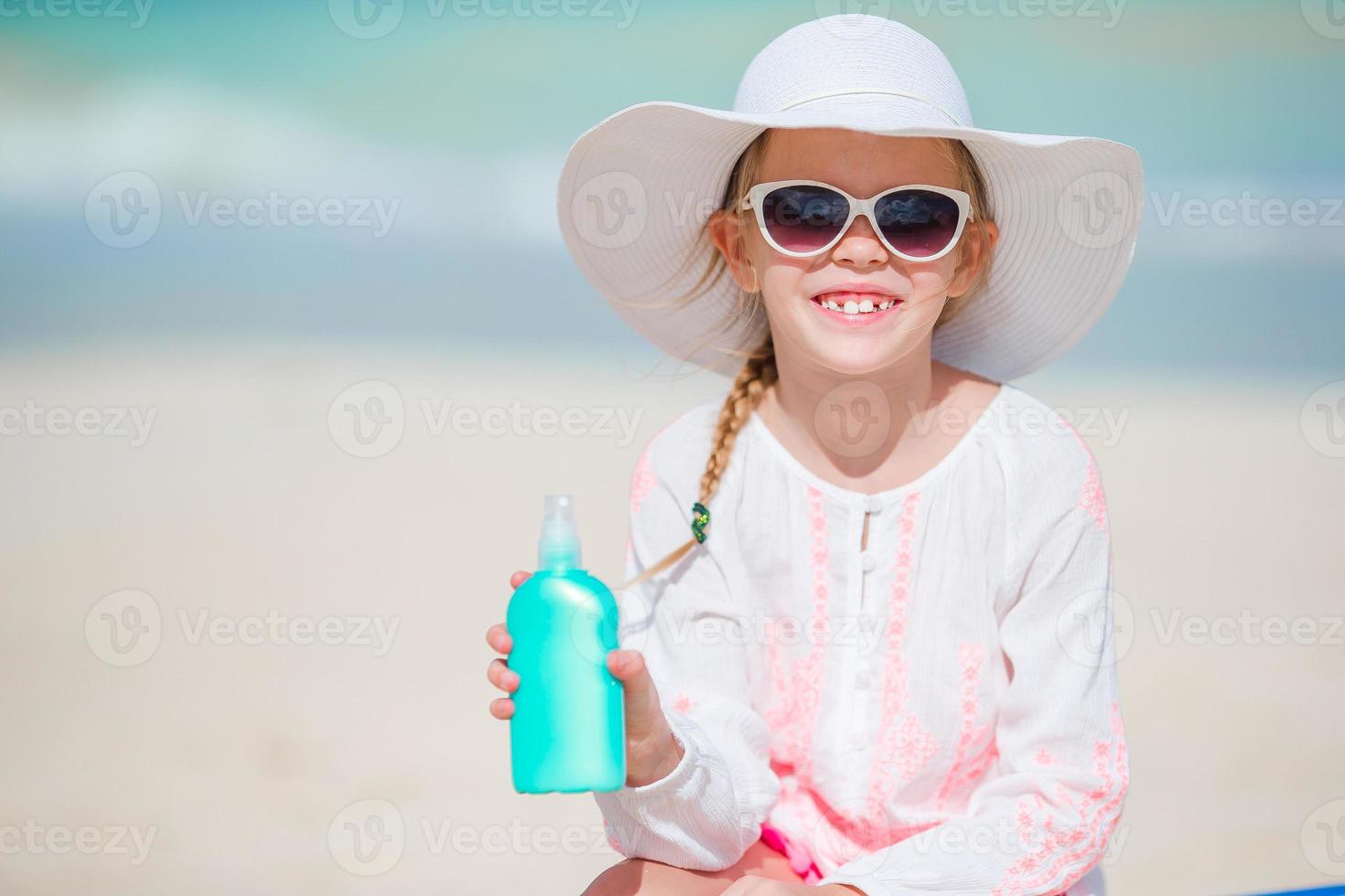 Little girl in hat with bottle of sun cream sitting at sunbed on tropical beach photo