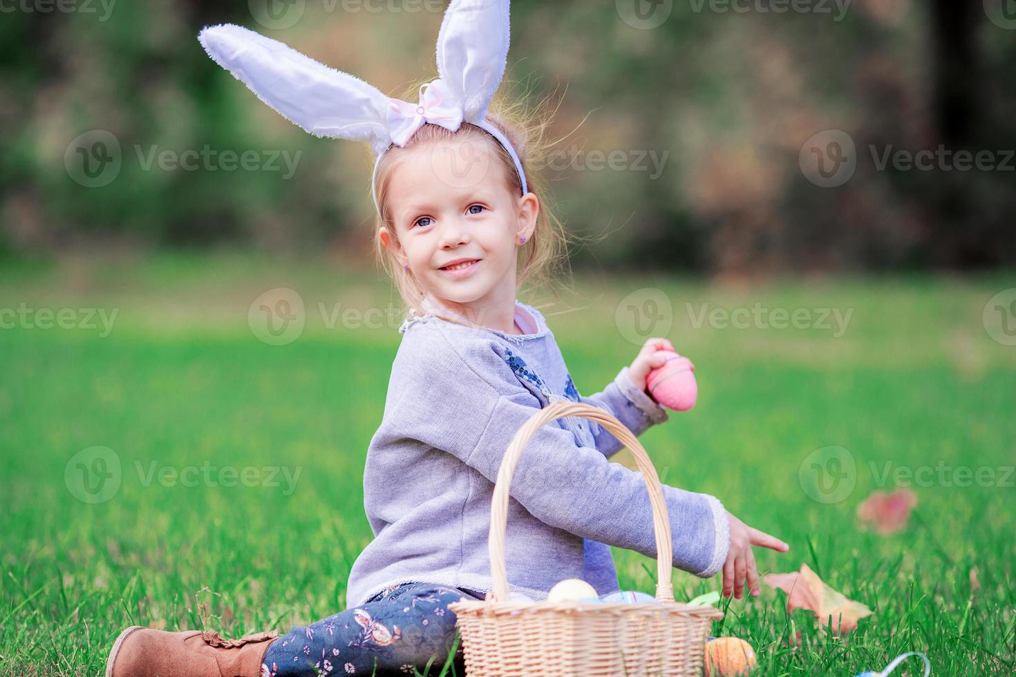 adorable niñita con orejas de conejo con una canasta llena de huevos de Pascua en primavera al aire libre foto