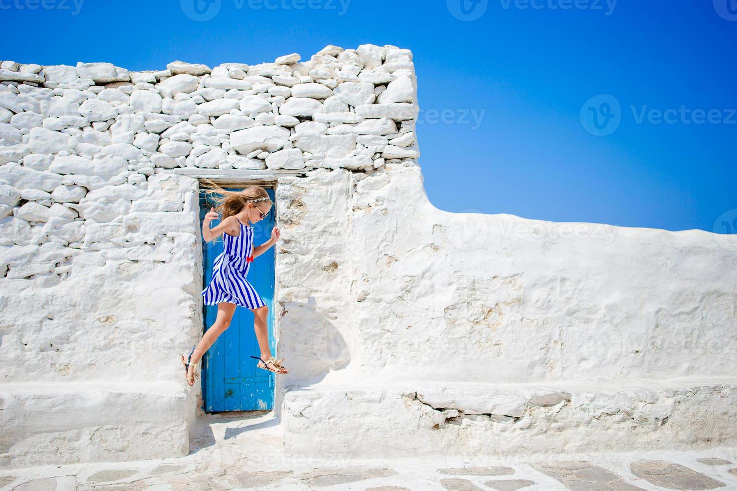 linda chica con vestido azul divirtiéndose al aire libre cerca de la iglesia paraportiani. niño en la calle del típico pueblo tradicional griego con paredes blancas y puertas coloridas en la isla de mykonos, en grecia foto