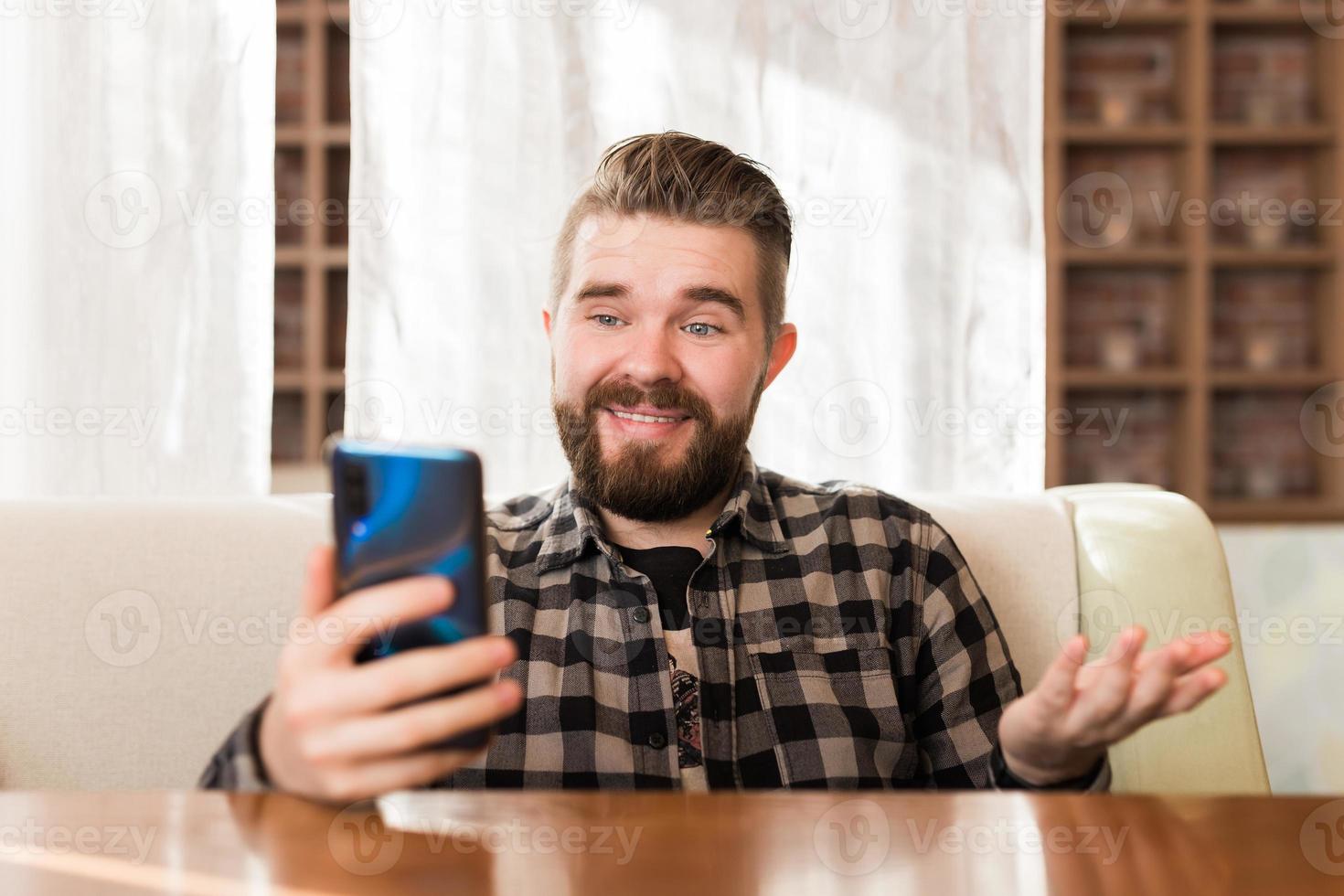 Happy young man sitting at coffee shot with earphones making video call to friend. Caucasian male having video chat on smart phone at cafe. photo