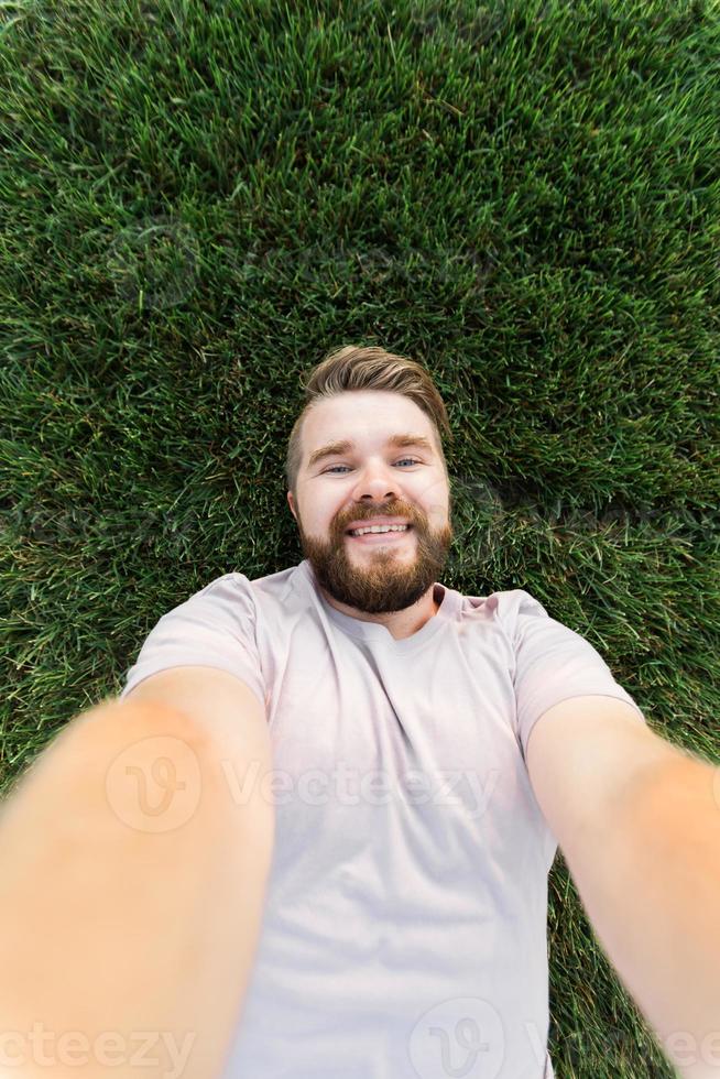 young man with beard and fashion hair style lying on grass taking selfie - holding smart phone or tablet and looking at camera. photo