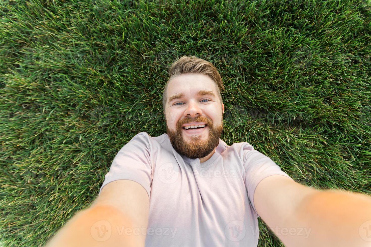 young man with beard and fashion hair style lying on grass taking selfie - holding smart phone or tablet and looking at camera. photo