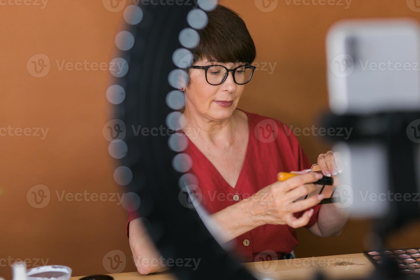 Middle-aged woman talking on cosmetics with makeup eye shadows and blush palette and brushes while recording her video. Mature female making video for her blog on cosmetics photo