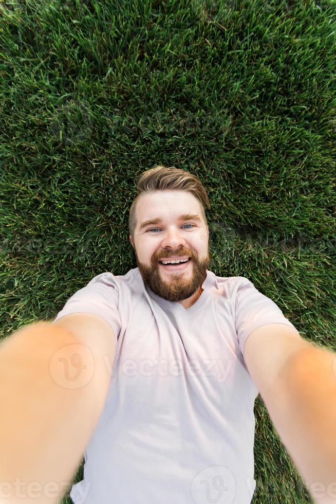 joven con barba y peinado de moda tirado en la hierba tomando selfie - sosteniendo un teléfono inteligente o una tableta y mirando la cámara. foto