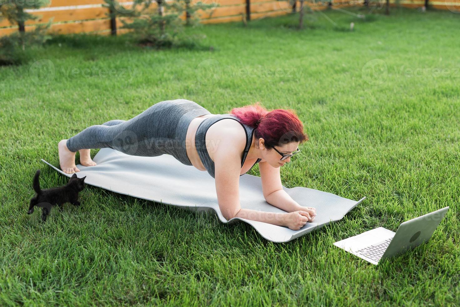 mujer joven de talla grande con top deportivo y polainas de pie en un tablón sobre una alfombra de yoga pasando tiempo en la hierba verde en el patio. gatito negro camina a su alrededor. concepto de bienestar y fitness foto