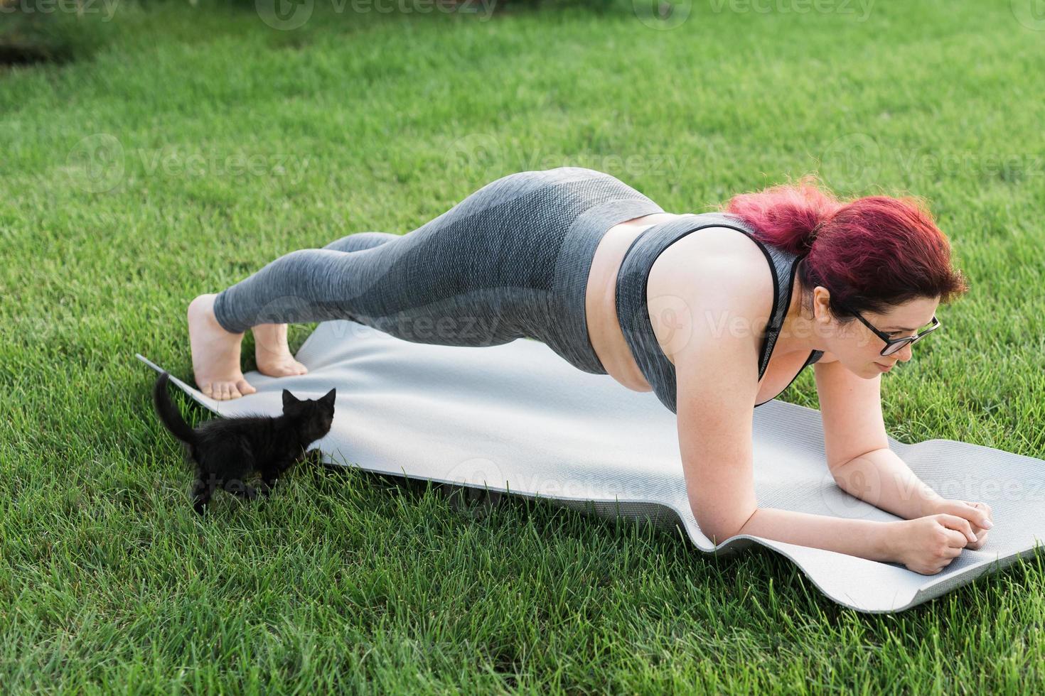 Young plus size woman in sporty top and leggings standing in plank on yoga mat spending time on green grass in yard. Black kitten walks around her. Well being and fitness concept photo
