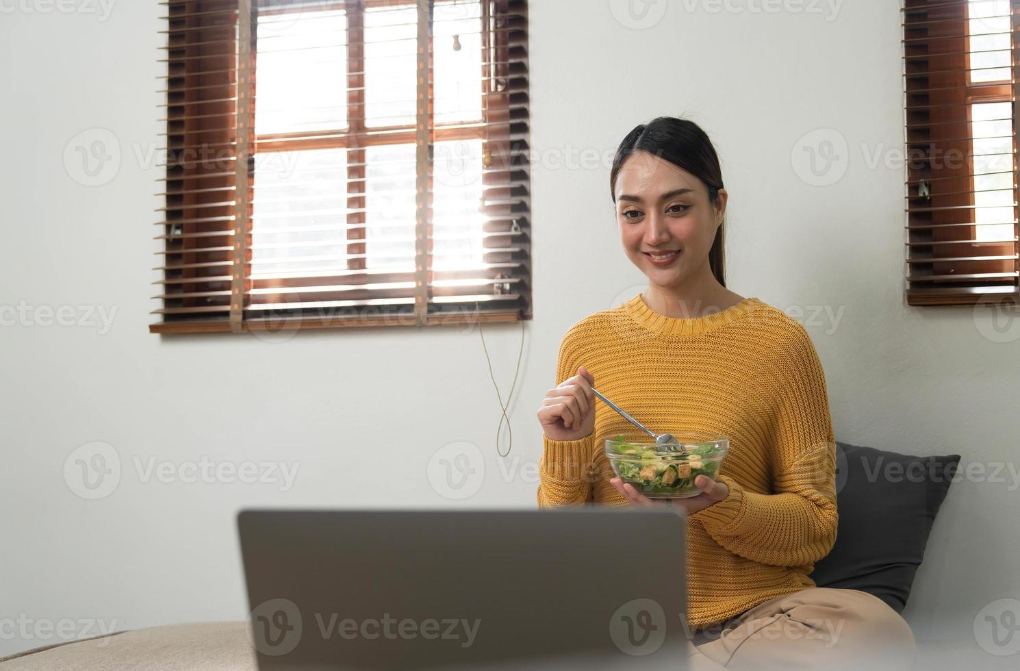 la gente se relaja en el hogar y el estilo de vida de bienestar. mujer asiática adulta joven comiendo ensalada y usando una computadora portátil para ver películas en línea en internet. foto