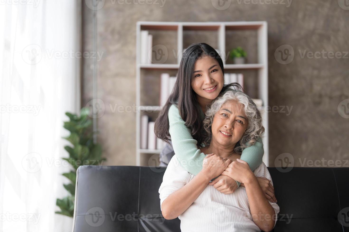 amorosa hija adulta abrazando a la madre mayor, parada detrás del sofá en casa, familia disfrutando juntos de un momento tierno, mujer joven y madre madura o abuela mirándose, dos generaciones foto