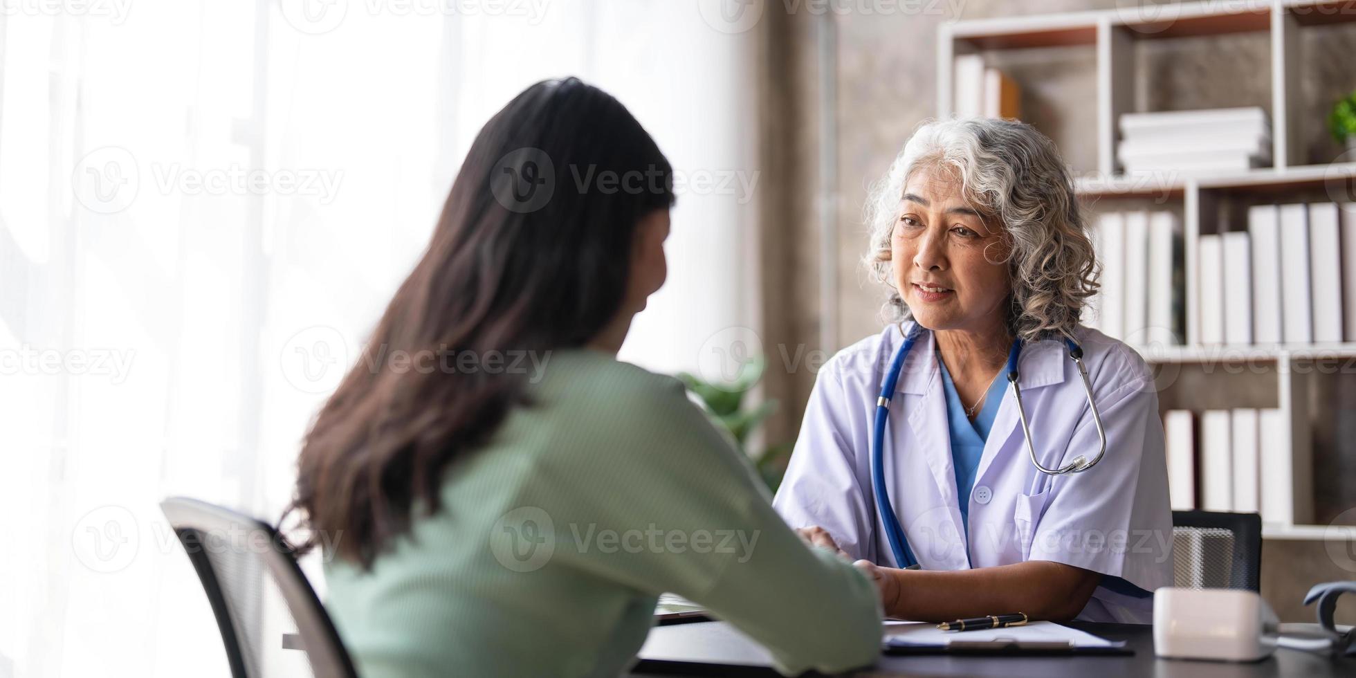 Woman senior doctor is Reading Medical History of Female Patient and Speaking with Her During Consultation in a Health Clinic. Physician in Lab Coat Sitting Behind a Laptop in Hospital Office. photo
